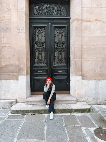Camila sentada diante de uma porta preta de ferro