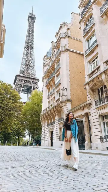 Camila caminhando por uma rua de prédios claros, com a Torre Eiffel ao fundo